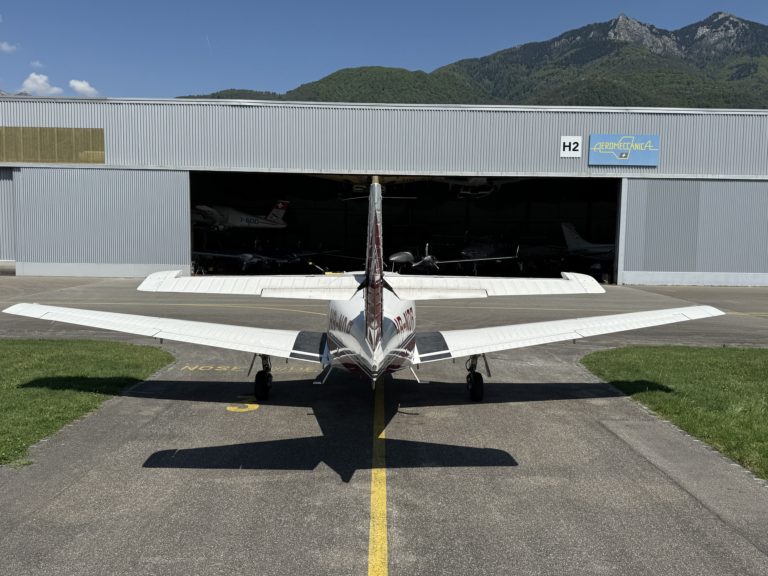 Small airplane parked outside hangar with mountain backdrop.